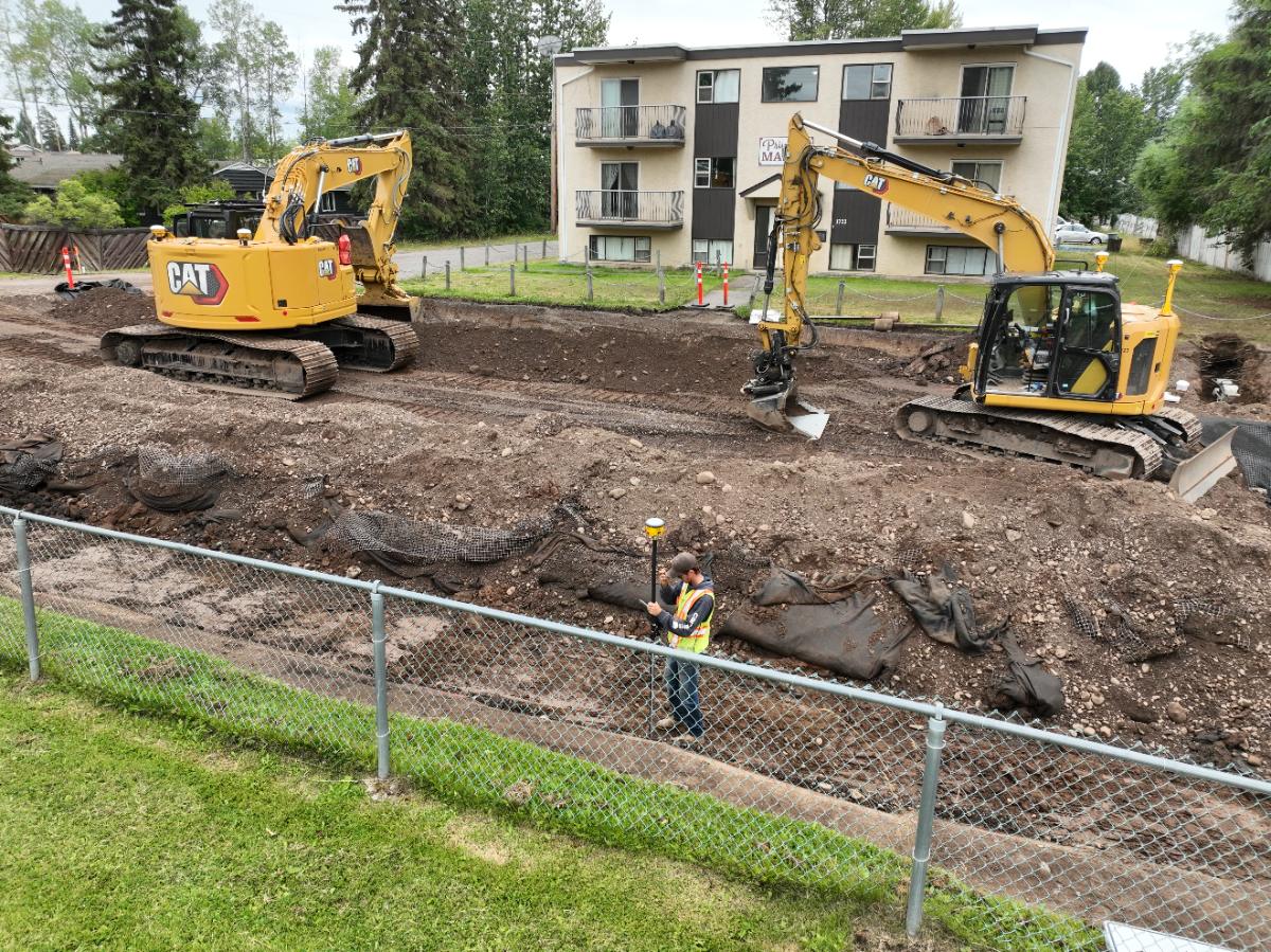 Two excavators and a worker with a GPS rover on a residential utility dig behind a chain-link fence